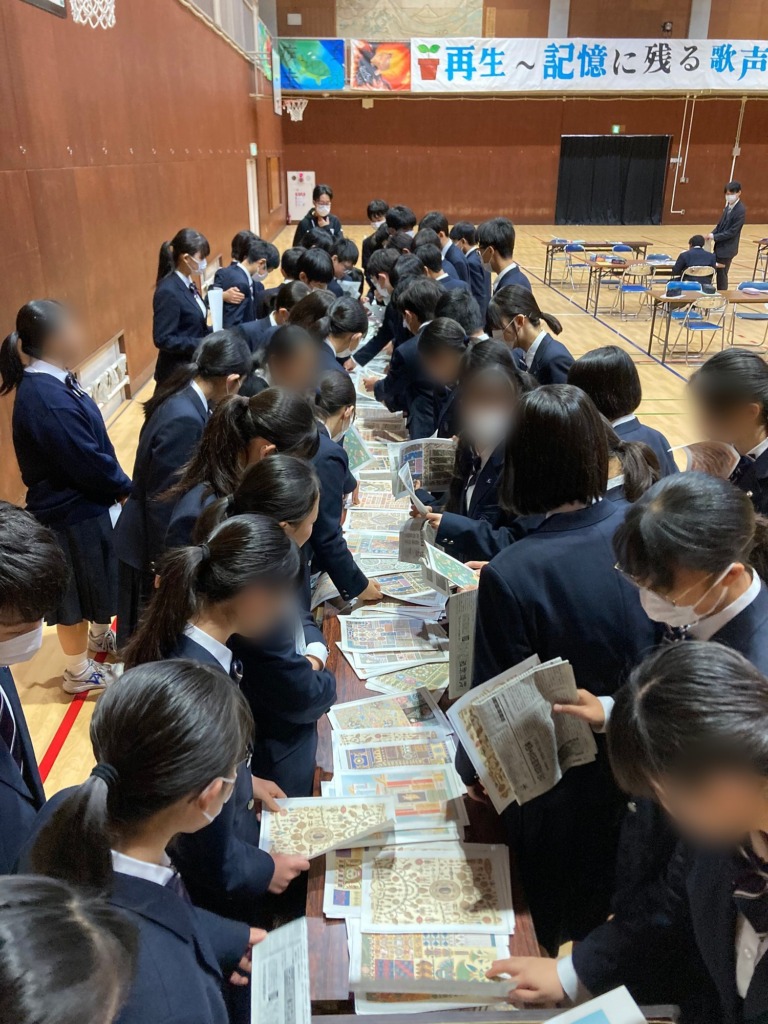 Students choosing collage materials laid out along a long table in a school gym